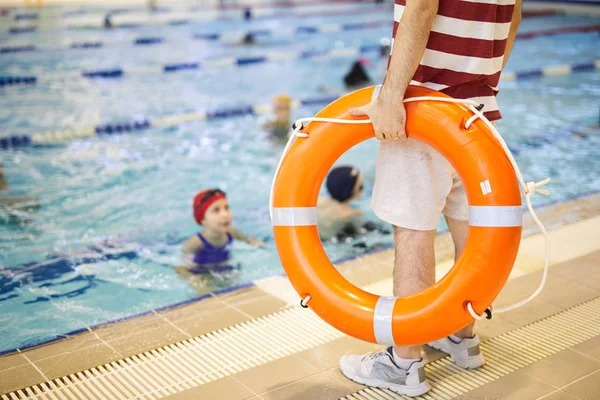 Close-up of swim trainer standing near the poolside and holding lifebuoy and training his class in the water