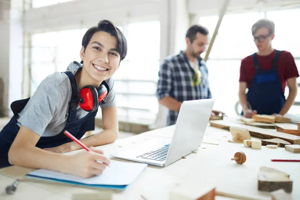 Cheerful excited young lady with ear protectors on neck leaning on table and smiling at camera while enjoying work in carpentry