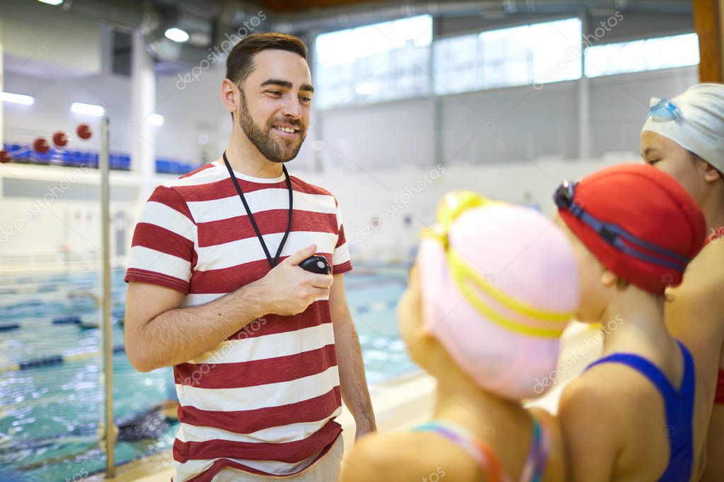 Young swim instructor standing with stopwatch and explaining children the rules of conduct in the pool