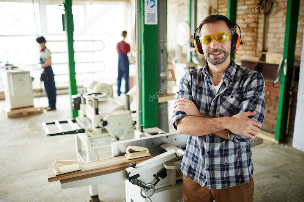 Cheerful handsome middle-aged carpenter in ear protectors and safety goggles standing against cutting machine and crossing arms on chest while smiling at camera