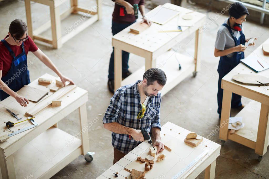 High angle view of busy carpenters standing at tables and working with wood while producing high quality toys, man using hand drill while making hole in wooden piece