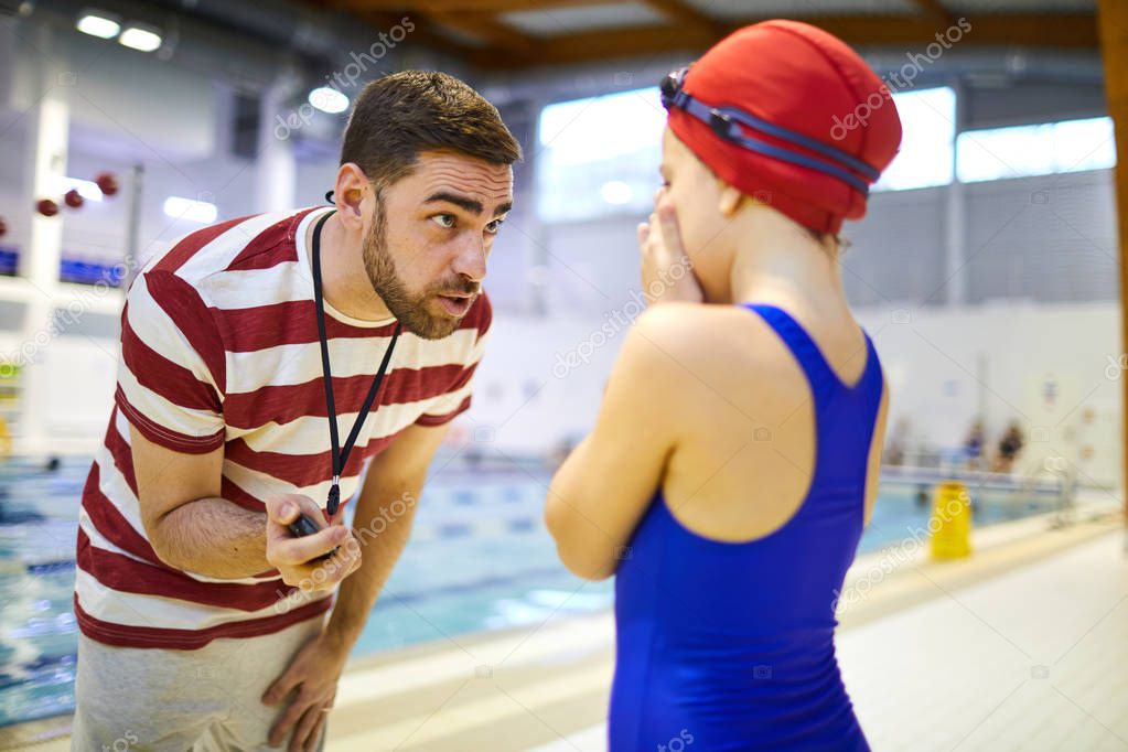 Serious swim instructor showing the results on his stopwatch to young athlete while she standing and closed her face with hands
