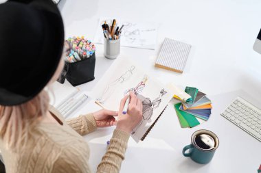 Şapkalı genç tasarımcının arka görünümü masada otururken ve kağıt üzerinde moda skeçleri çizerek