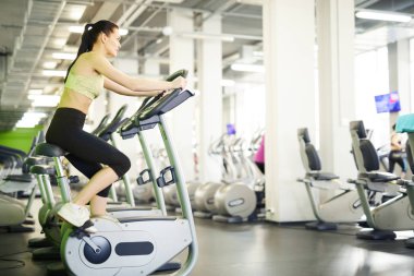 Spor giyinen genç aktif bir kadın spor bisikletlerinden birinde oturuyor ve spor salonunda egzersiz yapıyor.