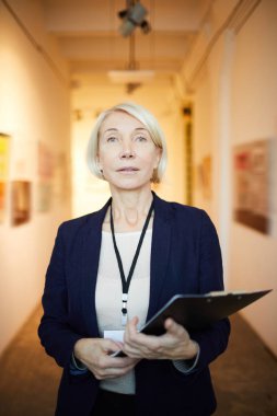 Waist up portrait of mature female manager holding clipboard posing in art gallery looking at camera clipart