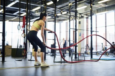 Dizleri bükülmüş, spor salonunda çapraz antrenman halatlarıyla egzersiz yapan genç aktif bir kadın.