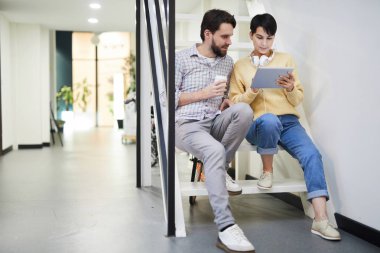 Merdiven üzerinde oturan ve proje üzerinde çalışırken tablet üzerinde çevrimiçi verileri analiz Casual giyim modern genç meslektaşları