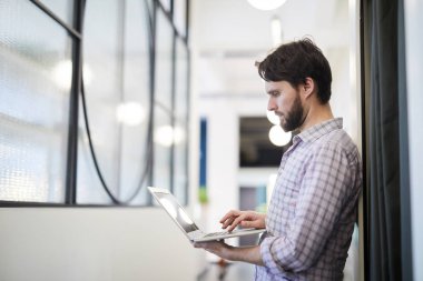 Ciddi konsantre Genç sakallı adam ofis koridorunda çevrimiçi veri ayakta odaklı ve internet verileri analiz ederken dizüstü bilgisayar kullanarak