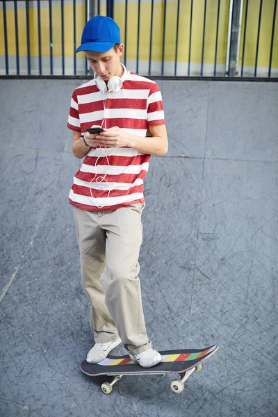 スケートボードの寒さの間にスマートフォンでカジュアルなTシャツ ズボン 野球帽とクロスシューズのメッセージングの男 — ストック写真