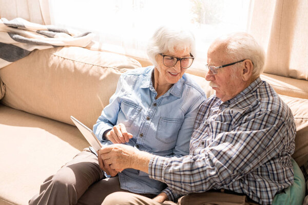 Portrait of modern senior couple using digital tablet sitting on couch at home in sunlight copy space