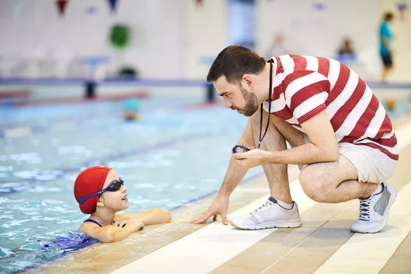 Serious swim trainer with stopwatch teaching little girl to swim in the pool and giving her some instructions