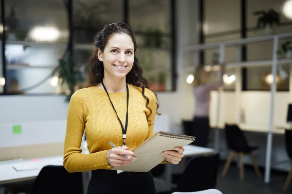 Smiling pretty young lady with badge on neck standing in modern open space office and holding sketchpad while looking at camera