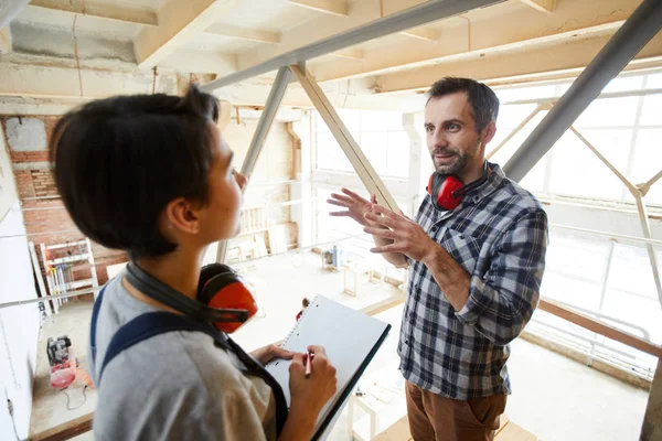 Content handsome bearded carpenter with ear protectors gesturing hands and explaining tasks to new employee while giving tour to her over carpentry