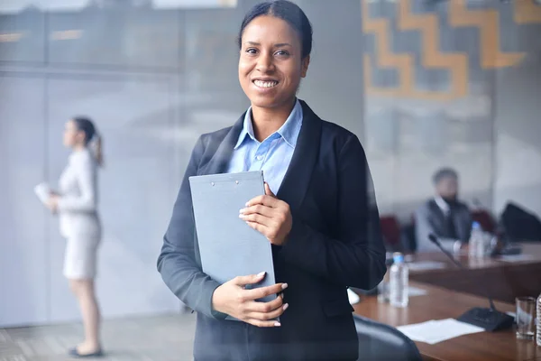 Young smiling businesswoman with clipboard looking at you in conference hall on background of colleagues