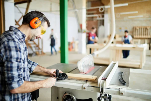 Serious concentrated handsome carpenter in ear protectors standing at workbench and adjusting wooden detail while preparing for cutting