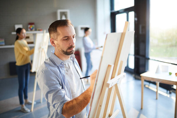 Serious young man in casualwear making sketch before painting on easel in contemporary studio of arts
