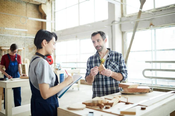 Content confident bearded carpenter in gray shirt standing at table with wooden detail and explaining intern how to work with wood, concentrated girl making notes during individual lecture