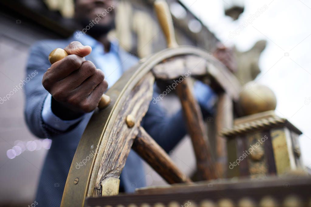 Hand of African-american business leader holding by large wooden sailing wheel while turning it