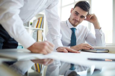 Young successful broker sitting by desk and looking at his business partner signing contract or financial paper