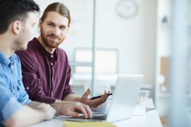 Young bearded creative designer looking at his colleague during explanation of his idea at meeting