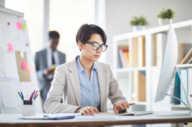 Young female broker in formalwear entering data and working with it in front of computer screen in office