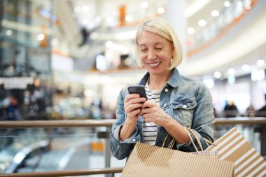 Alışveriş merkezi koridorunda duran ve alışveriş asistanı ile online sohbet ederken akıllı telefon sms mesajlaşma kısa sarı saçlı mutlu heyecanlı olgun kadın