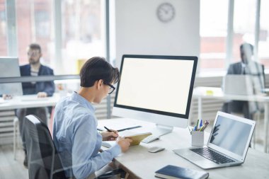 Young businesswoman in blue shirt making notes in notebook while sitting by desk in front of computer monitor