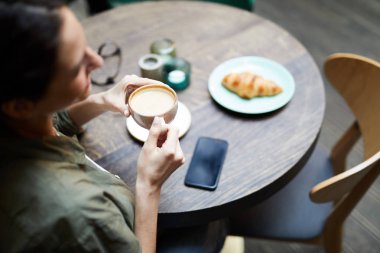 Genç bir kadın kahve dükkanında oturmuş kahvaltı ederken taze demlenmiş kahve içiyor.