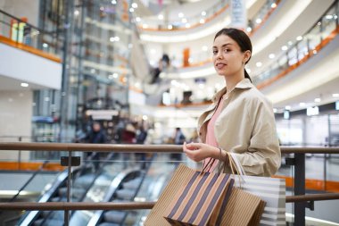 İçerik güzel genç bayan shopaholic midilli kuyruk ile bej ceket alışveriş merkezinde ayakta giyen ve kamera bakarken soyulmuş çanta tutarak