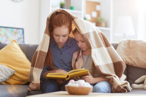 Portrait of two sisters reading book under blanket while sitting on sofa at home, copy space