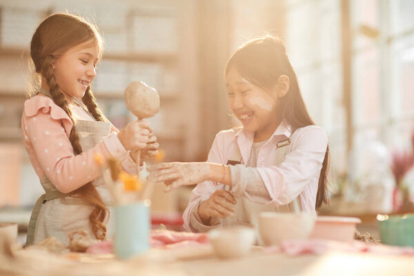 Portrait of two happy girls having fun in pottery art class lit by sunlight, copy space