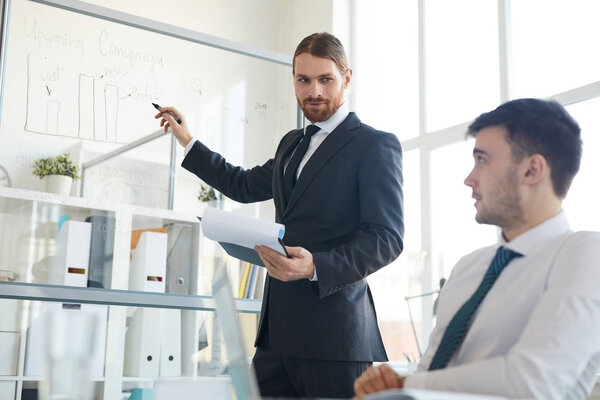 Young confident businessman in suit standing by whiteboad while pointing at chart during discussion with colleague