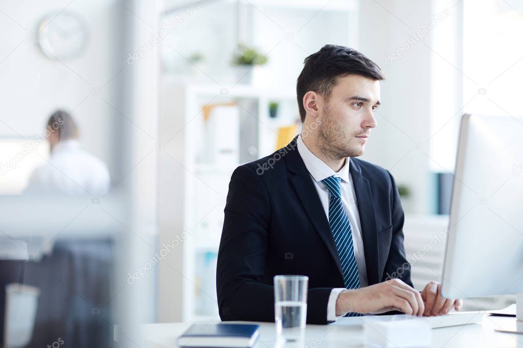Young elegant businessman in suit concentrating on network while sitting in front of computer screen