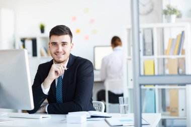 Online çalışma sırasında bilgisayar önünde işyeri tarafından oturan takım elbise neşeli ve başarılı genç komisyoncu