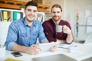 Young cheerful designers in casualwear sitting by desk in office or studio while looking through websites