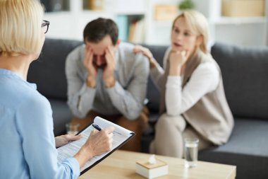 Sorunlu kocasının yanında oturan genç kadını dinlerken belgede notlar alan olgun profesyonel danışman