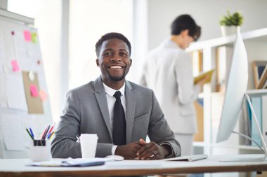 Young cheerful African-american businessman in formalwear sitting by table in front of camera during work in office