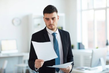 Young serious banker in formalwear reading contract or financial paper while working in office