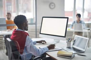 Young contemporary African-american office manager sitting by desk in front of computer screen and browsing in the net
