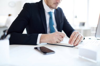 Young elegant office manager in formalwear making notes in notebook while sitting by his workplace