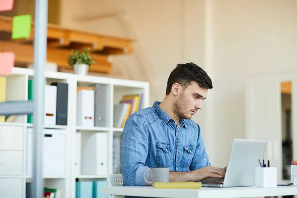 Jovem Trabalhador Escritório Sério Casualwear Sentado Mesa Frente Laptop Trabalhando — Fotografia de Stock