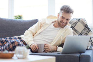 Kahve fincanı ile evde dinlenirken dizüstü ekrana bakan yakışıklı genç adamın portresi, kopya alanı