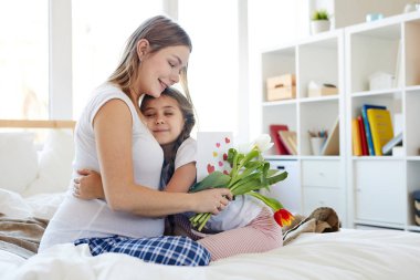 Hamile anne kucaklayan ve Anneler gününde ona çiçek veren sevimli küçük kız tam uzunlukta portre, her ikisi de sabah rahat yatakta oturan, kopya alanı