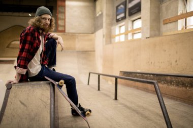 Aşırı parkta rampada oturan ve kameraya bakarak uzun saçlı patenci tam uzunlukta portre, kopya alanı