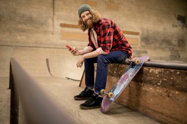 Uzun saçlı patencinin akıllı telefon kullanarak ve aşırı parkta rampada otururken kameraya gülümseyen tam uzunlukta portre, kopyalama alanı