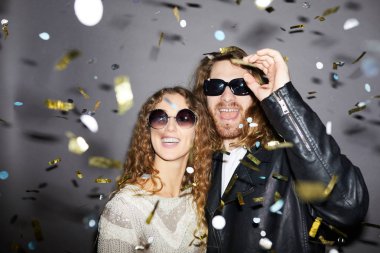 Altın konfeti altında duran ve partide eğlenmek güneş gözlüğü giyen kıvırcık saçlı neşeli heyecanlı hipster genç çift