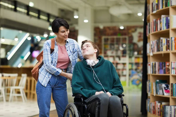 車椅子を押して 図書館で障害のある学生と話すランドセルを持つ肯定的な魅力的な若い女性 — ストック写真