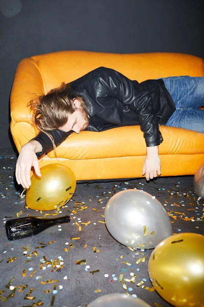Drunk young man in black jacket lying on sofa and sleeping after hilarious party in nightclub bottle, balloons and confetti on floor