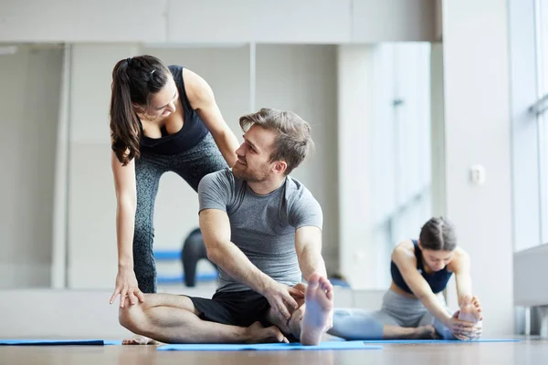 Content kind pretty yoga teacher talking to young man and adjusting his pose while he stretching leg at yoga class