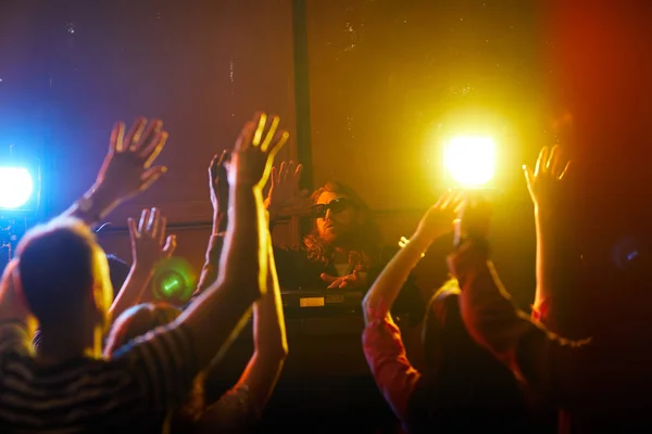 Brutal hipster young DJ in sunglasses twisting disc and waving hand together with crowd at party in nightclub